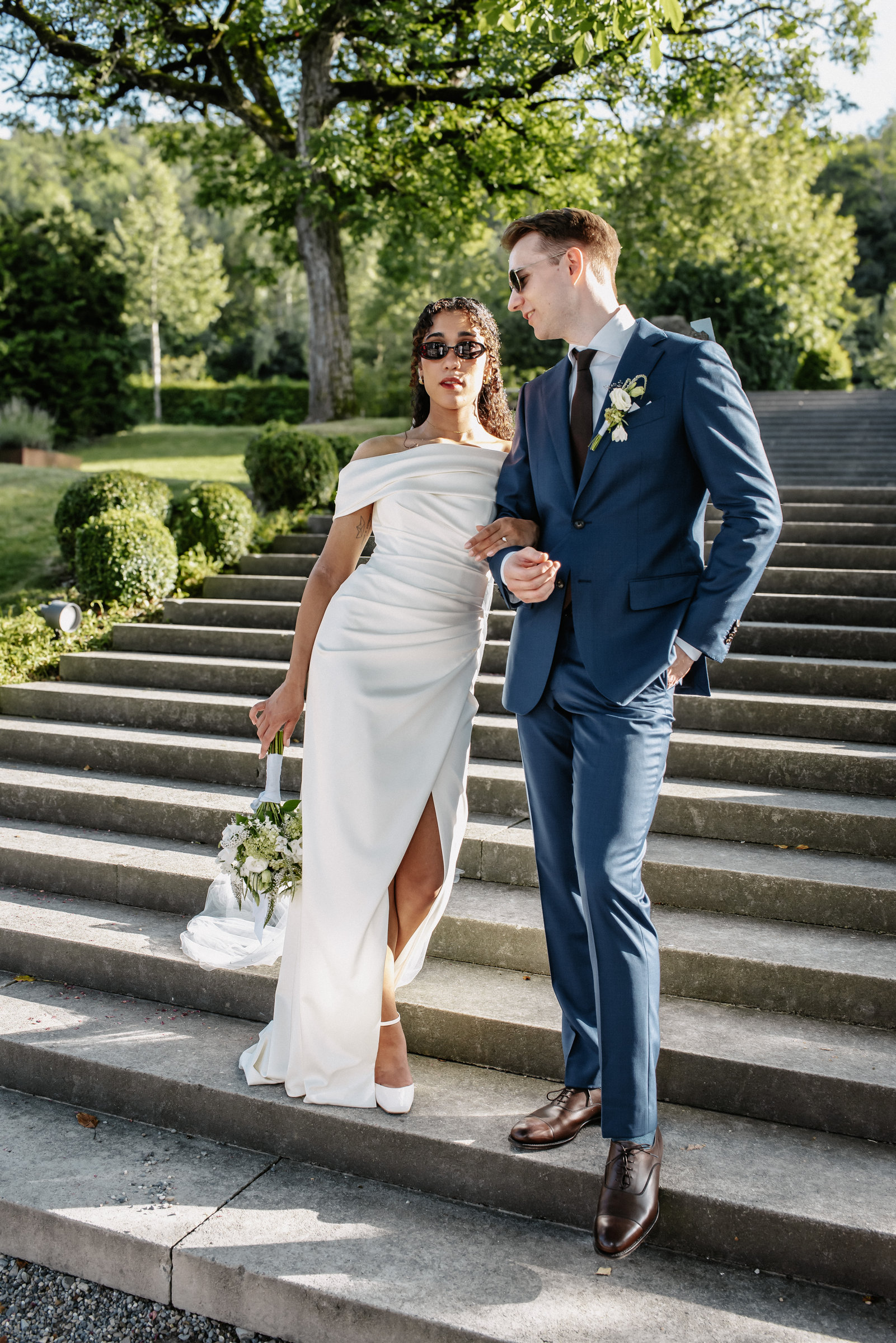 Elisa and Dominik in sunglasses descending sunlit stone steps — editorial wedding portrait