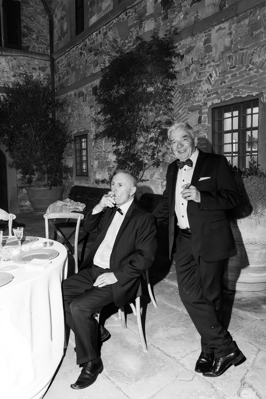Two guests in tuxedos sharing cigars in a castle courtyard, photographed unnoticed